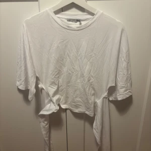 Vit knyt-topp från 157, storlek M - Enkel och stilren vit topp från 157 med korta ärmar och rund hals. Toppen har långa band i sidorna som kan knytas i midjan för en snygg detalj. Tillverkad i mjuk bomull, perfekt för en chill och avslappnad look.