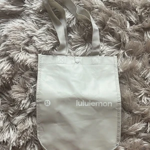Lululemon påse totebag - Snygg beige shoppingväska från Lululemon med vitt logotryck på framsidan. Väskan har långa handtag och är tillverkad i ett slitstarkt syntetmaterial. Perfekt storlek för att bära med sig allt man behöver under dagen.