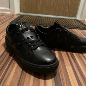 Svarta Valentino Rockstud sneakers - Snygga helsvarta Valentino Rockstud Untitled sneakers i skinn med ikoniska pyramidnitar på sidorna. Klassisk rund tå, svarta snören och diskret branding på plösen. Perfekta för dig som gillar en edgy och lyxig streetstil. Storlek 44. Men kan passa till 43.5
