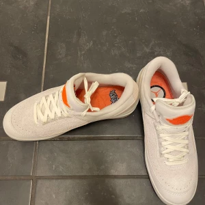 Nike Air Jordan 2 vita med orange detaljer - Nike Air Jordan 2 sneakers i vitt skinn och mocka med orange detaljer på plös och innersula. Kommer med extra skosnören i grönt och orange. Låg modell med klassisk Air Jordan-logga och cool text 'TEAM ONLY' på insidan av plösen.