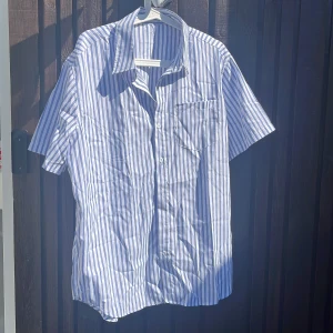 Randig kortärmad skjorta i bomull - Snygg skjorta med blå och vita ränder, klassisk krage och korta ärmar. Perfekt för varma dagar och enkel att matcha med jeans eller shorts. Skjortan har knappar framtill och en bröstficka. Tillverkad i mjuk bomull för en bekväm känsla.