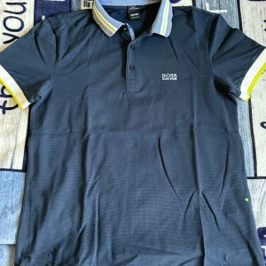 Mörkblå pikétröja från Hugo Boss - Snygg mörkblå pikétröja från Hugo Boss med slim fit-passform. Tröjan har klassisk krage med vita och gula detaljer samt korta ärmar med matchande kant. Tillverkad i lyxig bomull för en skön känsla. Boss-logga broderad på bröstet. priset ej hugget i sten vid snabbaffär 