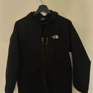 Svart vindjacka från The North Face - Svart vindjacka från The North Face med huva och hel dragkedja framtill. Jackan har en stilren look med logga på bröstet och praktiska fickor. Perfekt för dig som vill ha en enkel och funktionell jacka till vardags.