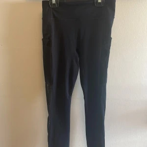 Svarta leggings från Fabletics - Svarta leggings från Fabletics Motion365 med hög midja och praktiska sidofickor. Tillverkade i stretchigt syntetmaterial som sitter tight och är perfekta för träning. Snygga detaljer längs benen ger extra stilpoäng.