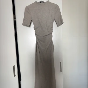 Beige klänning med rynkade detaljer - Säljer en beige långklänning med kort ärm och diskret rutigt mönster. Klänningen har rynkade detaljer i midjan som ger en snygg siluett. Materialet känns lätt och luftigt, perfekt för varmare dagar. Enkel och stilren design som är lätt att matcha.