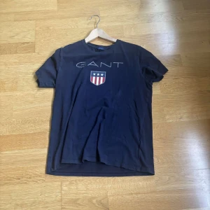 Mörkblå GANT t-shirt med tryck - Snygg mörkblå t-shirt från GANT med stort tryck framtill och sköld med stjärnor och ränder. Klassisk rund hals och korta ärmar. Tillverkad i mjuk bomull som känns skön mot huden. Perfekt för en avslappnad och stilren look.