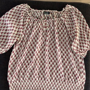 Blommig blus från ONLY med pärlknappar - Supersöt, transparent blommig blus från ONLY i vitt med röda och svarta blommor. Blusen har söta ärmar, smockdetaljer vid hals och nederkant samt pärlknappar framtill. Tunt och luftigt material som passar perfekt till vår och sommar. Väldigt fin över ett vitt linne. Står S men passar bäst till M.