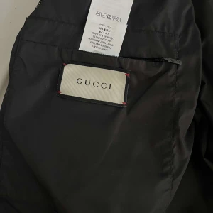 Svart bomberjacka från Gucci - Svart bomberjacka från Gucci med stort färgglatt logotryck på ryggen och klassisk ribbad krage i rött, vitt och blått. Jackan har dragkedja framtill, två fickor med dragkedja och elastiska muddar vid ärmslut. Tillverkad i Italien och i polyester.