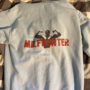 Ljusblå Gildan hoodie med tryck revinite og  - Ljusblå hoodie från Gildan med stor ficka framtill och huva. På ryggen finns ett tryck med texten 'MILFHUNTER' och en flexande siluett. Tröjan är i mjuk bomullsblandning och har en avslappnad passform, perfekt för chill dagar.