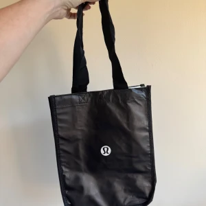 Svart shoppingväska från Lululemon - Säljer en svart och vit shoppingväska från Lululemon med deras logga på framsidan och tryckt text på sidan.