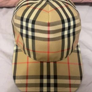Burberry keps - Snygg keps från Burberry med klassiskt rutigt mönster i beige, svart, vitt och rött. Justerbar baktill med svart spänne och tydlig Burberry London England-logga. Perfekt accessoar för att lyfta din outfit med en lyxig touch.