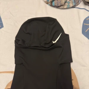 Svart Nike Pro balaclava - Svart balaclava från Nike Pro med vit swoosh-logga på sidan. Tillverkad i stretchigt syntetmaterial som andas, perfekt för träning utomhus. Snygg, tight passform och diskreta sömmar för en clean look. Märke på baksidan. Passar dig som vill ha sportig stil.