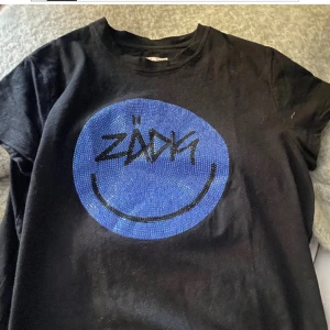 Svart Zadig & Voltaire t-shirt med blått tryck - Cool svart t-shirt från Zadig & Voltaire med ett stort blått smiley-tryck. Köpt i Paris i butiken för ca 1500kr.