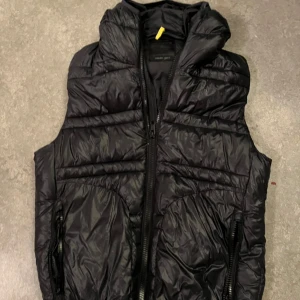 Svart dunväst från Replay - Snygg svart dunväst från Replay med quiltad design och hög krage. Västen har dragkedja framtill och två stora fickor med dragkedja. Tillverkad i glansig polyester som ger en modern look. Perfekt att slänga över en hoodie för extra värme.