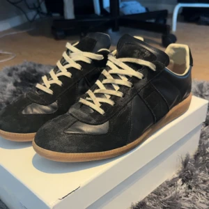 Maison Margiela svarta sneakers i skinn - Maison Margiela sneakers i svart skinn och mocka med vita snören och klassisk gummisula. Snygg retrostil med rund tå och diskreta paneler. Perfekta för dig som gillar stilrena och exklusiva sneakers med en lyxig känsla.
