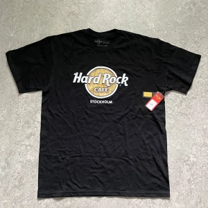Hard Rock Cafe svart t-shirt Stockholm - Svart t-shirt från Hard Rock Cafe med klassiskt tryck i vitt och guld på bröstet där det står Stockholm. Skön passform, rund hals och korta ärmar. Perfekt för dig som gillar ikoniska bandtröjor och vill sticka ut med en cool look.