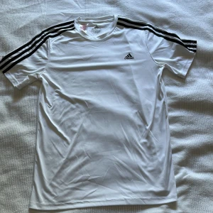Vit Adidas t-shirt med svarta ränder - Snygg vit t-shirt från Adidas med klassiska svarta ränder längs axlarna och Adidas-logga på bröstet. Tillverkad i ett lätt och sportigt polyestermaterial, perfekt för träning eller chill. Kortärmad och med rund hals.
