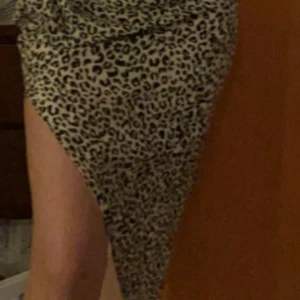 Bershka  - Snygg långkjol med leopardmönster i svart och beige. Kjolen har en asymmetrisk form med hög slits framtill och draperad detalj i sidan. Resår i midjan för skön passform och dragkedja. Perfekt statement-plagg för dig som vill sticka ut.