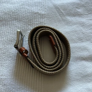 Beigefärgat repbälte från Jack & Jones - Säljer ett snyggt beigefärgat bälte från Jack & Jones med repstruktur och detaljer i brunt skinn. Bältet har en klassisk silverfärgad metallspänne och passar perfekt för att ge outfiten en avslappnad men stilren touch.