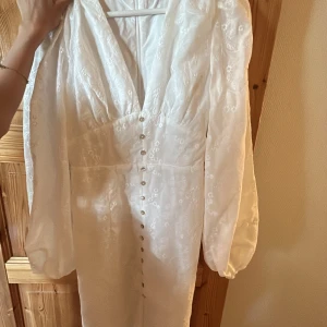 Vit långklänning med puffärm och slits -  Vitlångklänning med v-ringning, puffiga långärmar och knappar framtill. Klänningen har broderade blommor i tyget, markerad midja och en slits framtill. 