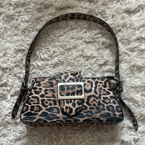 Leopardmönstrad axelväska med spänne - Cool axelväska med leopardmönster och bred axelrem. Väskan har ett stort silverfärgat spänne framtill och dragkedja upptill. Perfekt för dig som vill sticka ut med en edgy accessoar. Väskan är köpt second hand men var helt oanvänd, har nu använts fåtal gånger. 