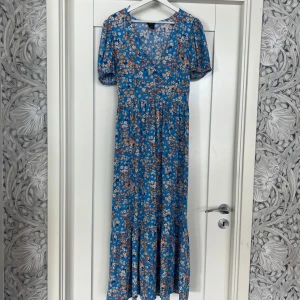 Blommig blå långklänning Melissa - Supersöt långklänning från Lindex i blått med små vita och orange blommor. Klänningen har v-ringning, korta puffärmar och volang nertill. Perfekt för sommarens alla tillfällen och ger en fräsch och färgglad vibe. Aldrig använd bara provad! 