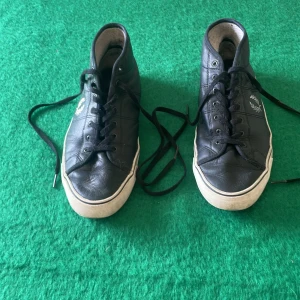 Svarta Fred Perry sneakers i skinn - Svarta sneakers från Fred Perry i skinn med högt skaft och klassisk vit logga på sidan. Skorna har svarta snören, vit sula och rund tå. Perfekta för en stilren och sportig look.