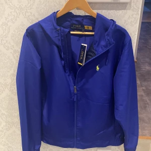 Blå vindjacka från Polo Ralph Lauren - Snygg blå vindjacka från Polo Ralph Lauren med huva och dragkedja framtill. Jackan har klassisk gul broderad logga på bröstet och justerbar snörning i huvan. Perfekt för dig som gillar stilren och sportig look. Storlek L men passar M