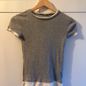 Grå ribbad kortärmad topp - En enkel och stilren grå ribbad topp med korta ärmar och vita detaljer vid halsringning, ärmslut och nederkant. Toppen har en tight passform och är perfekt för en clean och avslappnad look. 