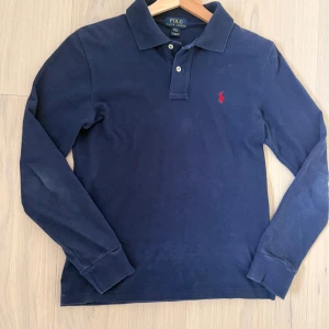 Marinblå pikétröja från Polo Ralph Lauren - Klassisk marinblå långärmad pikétröja från Polo Ralph Lauren med röd broderad logga på bröstet. Tröjan har krage och knäppning med två knappar framtill. Perfekt för en stilren och tidlös look. Storlek xs=14-16 år