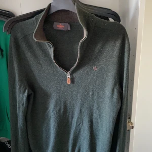Mörkgrön halvzip-tröja från Morris - Säljer en mörkgrön stickad tröja från Morris med halv dragkedja och liten broderad logga på bröstet. Tröjan har lång ärm och ribbade muddar. Perfekt för lager-på-lager under kyliga dagar.