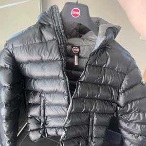 Svart dunjacka från Colmar - Säljer en svart dunjacka från Colmar med huva och dragkedja framtill. Jackan har en klassisk pufferstil med horisontella sömmar och två fickor med dragkedja. Perfekt för kyliga dagar och enkel att matcha med olika outfits. Nypris 5500kr, mitt pris 1200kr! 