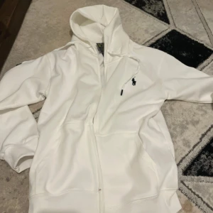 Vit hoodie från Polo Ralph Lauren - Säljer en fräsch vit hoodie från Polo Ralph Lauren med dragkedja och klassisk blå logga på bröstet. Tröjan har huva med snörning och två fickor framtill. Perfekt för en clean och stilren look.