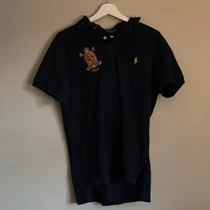 Marinblå pikétröja från Polo Ralph Lauren - Klassisk svart pikétröja från Polo Ralph Lauren med broderad logga på bröstet och snygg detalj med emblem. Tröjan har korta ärmar och krage med knappar. Perfekt för sommaren.