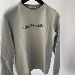 Ljusgrå sweatshirt från Calvin Klein Jeans - Säljer en stilren ljusgrå sweatshirt från Calvin Klein Jeans med svart logga på bröstet och ärmen. Tröjan har rund halsringning och ribbade muddar vid ärmslut och nederkant. Perfekt för en avslappnad look.