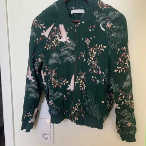 Grön bomberjacka med fågel- och blommönster från Promod - Säljer en grön bomberjacka från Promod med dragkedja. Jackan har ett detaljerat mönster med vita fåglar och blommor i rosa och beige toner.