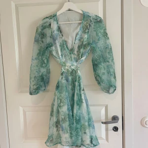 Blommig grön och blå omlottklänning - Säljer en superfin blommig klänning i gröna och blå toner med v-ringning och långärmade, transparenta ärmar. Klänningen har omlottdetalj i midjan och öppen rygg, perfekt för sommarens tillfällen. PRIS GÅR ATT DISKUTERAS 