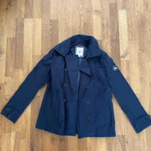 Mörkblå skepparkavaj  - Stilren mörkblå skepparkavaj från key west med dubbelknäppning och klassisk krage. Jackan har två snedställda fickor framtill och en patch med logga på ena ärmen. Perfekt för en snygg och tidlös look.