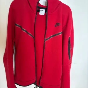 Röd hoodie från Nike med dragkedja - Röd hoodie från Nike med svart logga på bröstet och svarta detaljer längs dragkedjan och ärmen. Tröjan har huva, hel dragkedja och en ficka med dragkedja på ena ärmen. Perfekt för en sportig look. Tröjan är i bra skicka, men dragkedjan är lite sliten som man kan se på bilden 