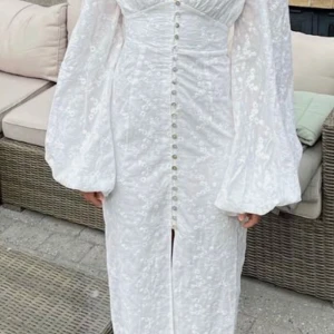 Adooreklänning  - Superfin vit långklänning med v-ringning, puffiga långärmar och dekorativa knappar framtill. Klänningen har ett subtilt blommigt mönster och markerad midja. Perfekt för sommarens speciella tillfällen.