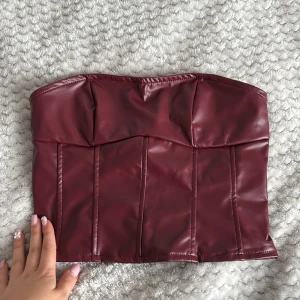 Vinröd korsett-topp i skinnimitation - Säljer en vinröd korsett-topp i skinnimitation med snörning i ryggen. Toppen är axelbandslös och har markerade sömmar framtill för en snygg passform. Perfekt för att lyfta vilken outfit som helst! Den är oanvänd och säljs just för att den ej kommer till användning. Från shein