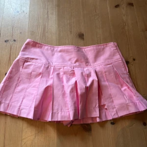 Rosa plisserad kortkjol - Säljer en söt rosa plisserad kortkjol med bred linning. Kjolen har klassiska veck och är perfekt för dig som gillar en sportig och trendig stil. Den har shorts under! Storlek L men man kan vika ner den till strl M