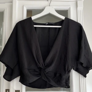 Svart omlottopp från Zara - Köp via instabox!!! Säljer en svart omlottopp från Zara med korta ärmar och knytdetalj framtill. Toppen har en djup v-ringning och är croppad.