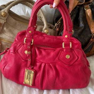 Röd Q Classic Baby Groove väska! - Snygg röd handväska i bra skick från Marc by Marc Jacobs i äkta läder med gulddetaljer och dubbla handtag. Finns lite slitage på fötterna på undersidan, men inget tydligt alls. Inga repor eller hål på väskans läder. Lite slitet vid handtagen som syns på bilden. Välbevarad vintage modell! Perfekt för att ge en stilren touch till din outfit. Skriv privat för fler bilder. Mått 34x18x12 (undersida). Kan hämtas hemma hos mig i Nacka eller skickas men kan tyvärr inte mötas🤍