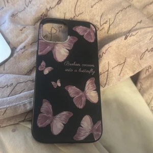 Svart mobilskal med lila fjärilar (iPhone 15) - Säljer ett svart mobilskal dekorerat med flera lila fjärilar och texten 'Broken cocoon into a butterfly' på baksidan. Skalet har en matt finish och är gjort i ett flexibelt material som skyddar mobilen.(iPhone 15)