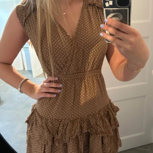 Prickig klänning med volanger från Designers Remix Girls - Skön söt prickig klänning från designers remix girls!! Klänningen har storleken ”14 år” och kan knäppas vid bysten. V ringning, brun med vita prickar, kortärmad och volangdetaljer. Passar bra till nästan alla årstider!! Säljer då den är för liten (klänningen är ostruken på bilderna)