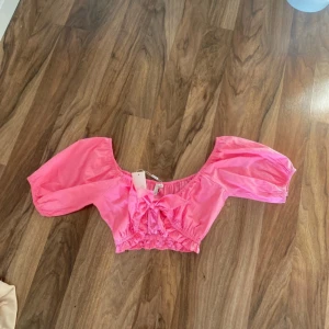 Rosa croppad blus med volang och rosett - Säljer en söt rosa croppad blus med volangdetaljer och rosett framtill. Blusen har korta puffärmar och resår i midjan för en bekväm passform. Perfekt för varma dagar och en trendig look!