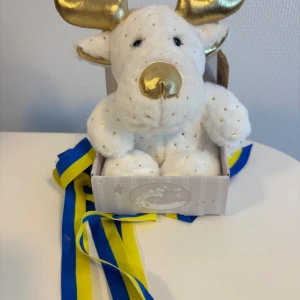 Vit gosedjursälg med gulddetaljer för studenten - Supergullig vit älg i mjukt plyschmaterial med små guldfärgade prickar och horn samt nos i metallicguld. Sitter i en ljus kartong med stjärnmönster. Perfekt som present, och har blågula band så du kan ge studenten att hänga runt halsen. 