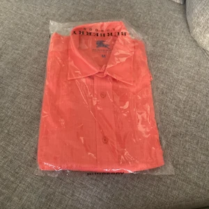 Korallrosa skjorta från Burberry - Säljer en snygg korallrosa skjorta från Burberry i storlek M. Skjortan har klassisk krage och knäppning framtill. Perfekt för dig som vill ha en stilren och färgstark look. Skjortan är i ett lätt material och levereras i originalförpackning.
