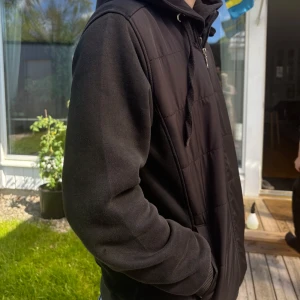 Svart hoodiejacka från Armani - Svart hoodiejacka från Armani med broderad logga på bröstet och rund EA-detalj på ärmen. Jackan har dragkedja framtill, huva med snörning och quiltade partier på framsidan. Perfekt för en stilren och sportig look.
